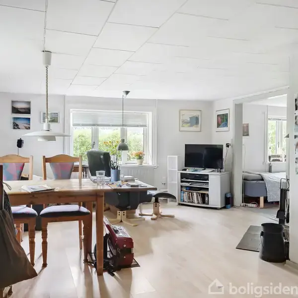 Et lyst spisestueområde med et træbord og stole, beliggende ved store vinduer. I baggrunden ses en stue med et tv, bøger og åbning til et soveværelse. Væggene har billeder og malerier.