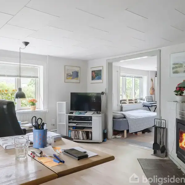 En stue med brændeovn, hvor en flamme brænder. Et spisebord med skriveartikler står nær et stort vindue. I baggrunden ses et tilstødende rum med en seng.