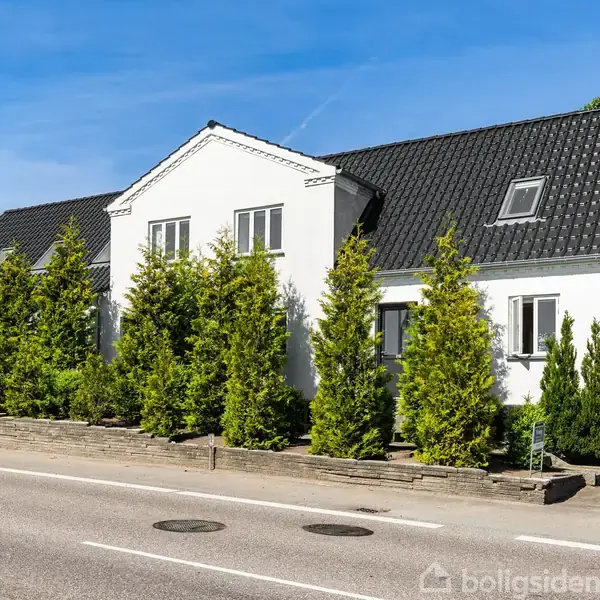 Hvidt hus med sort tag står langs en vej, omgivet af grønne buske. Klar himmel i baggrunden.