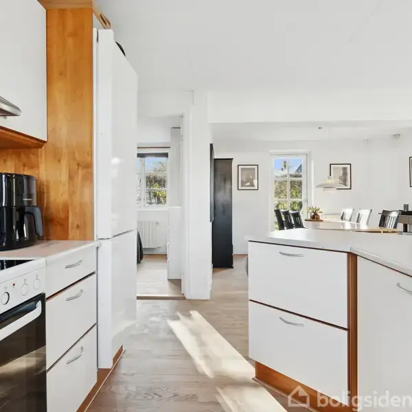 Køkken med hvide skabe og sort komfur, indeholder en kaffemaskine; det åbner op til en lys spisestue med trægulv og store vinduer, der slipper naturligt lys ind.