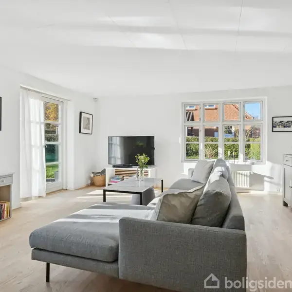 Grå sofa står i lys stue med parketgulv. Tv bord og skærm i hjørne, omgivet af vinduer med udsigt til grøn have. Moderne og minimalistisk indretning.