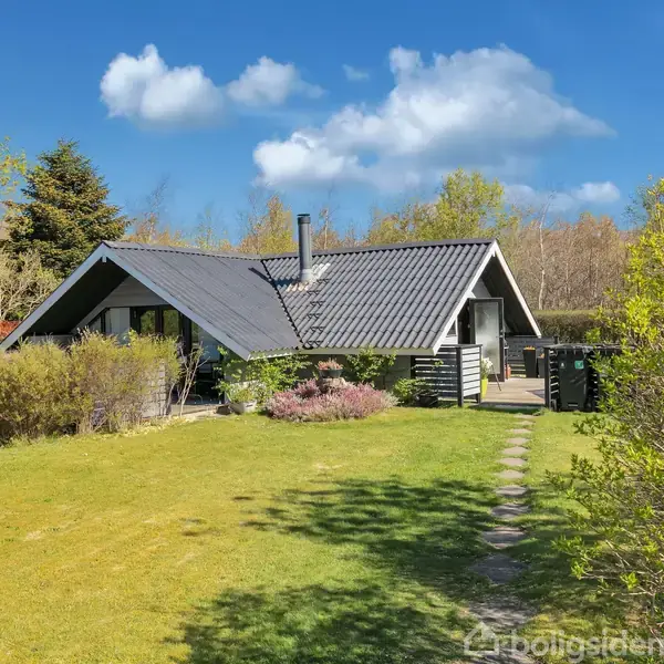 En sortmalet sommerhus står stille omgivet af grønne buske og en velplejet græsplæne med en flisegang. Blå himmel og træer i baggrunden skaber en fredelig atmosfære.