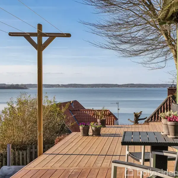 Træterrasse med bord og stole vender mod havudsigt under en klar blå himmel, omgivet af huse og træer. Potteplanter dekorerer terrassen, som har et højt, træbeklædt hegn i højre side.