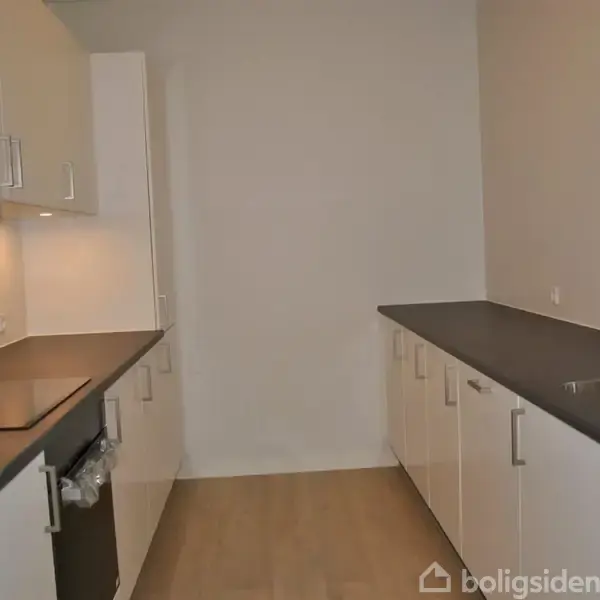 Et køkken med hvide skabe og sorte bordplader, der indrammer rummet; komfur og vask på hver side, med trælaminatgulv omgivet af lyse vægge.