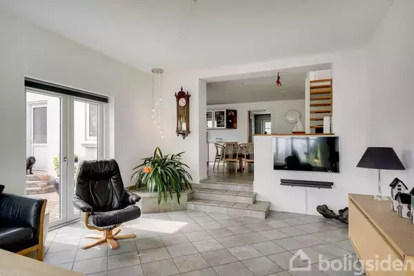 En stue med en sort lænestol og en stor plante placeret på flisegulvet. En vægmonteret fladskærm hænger ved siden af indgangen til spisestuen, der er på et hævet niveau.