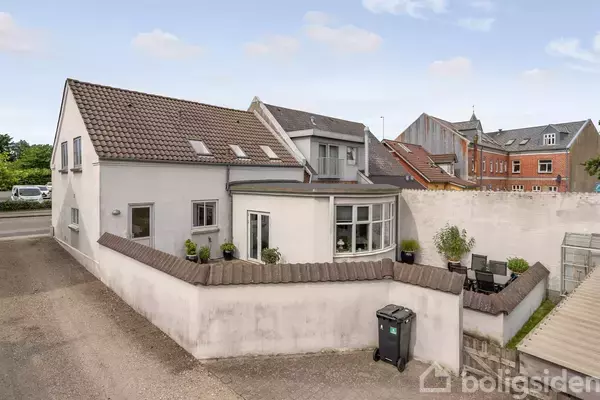 Et hvidt hus med skråtag og kviste står ved en vej. Forhaven har planter og en udendørs siddeplads, omgivet af en lav mur. Bagved ses andre bygninger.