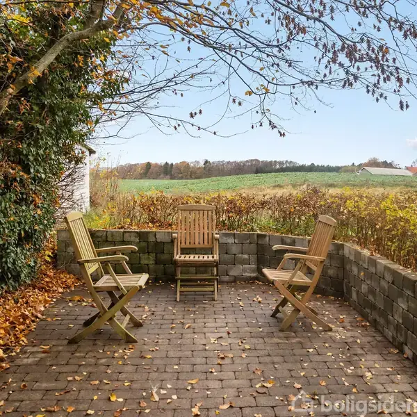 Tre træstole står stille på en brolagt terrasse omgivet af en murstensmur og efterårsblade. I baggrunden ses en udsigt over grønne marker og fjerne træer.