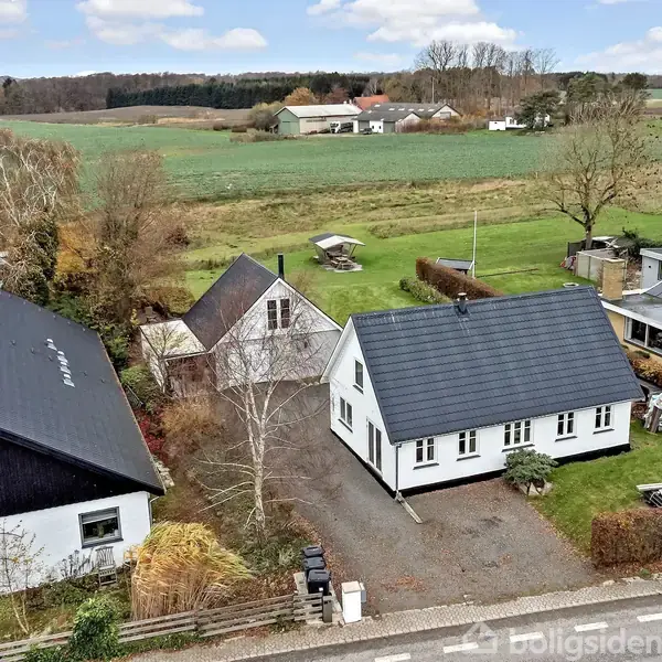 Et hvidt hus med sort tag står på en grusvej omgivet af hække og tæt på andre huse. I baggrunden ses store grønne marker og skovbryn under en skyet himmel.