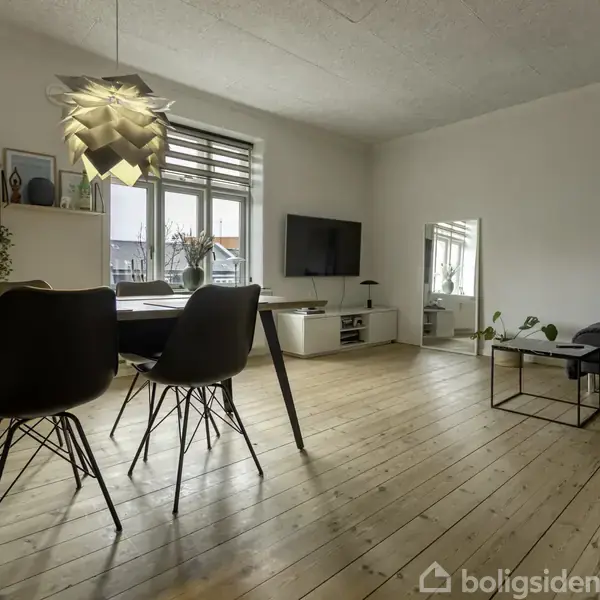 Et moderne stueinteriør med et spisebord og stole i forgrunden. En lampe hænger over bordet. Rummet har en sofa, et TV på væggen og træplankegulv.
