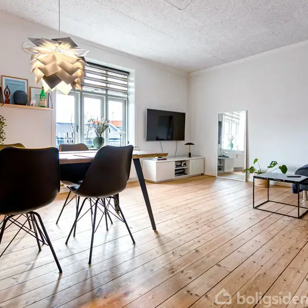 Et spisebord med sorte stole står på et trægulv. Over bordet hænger en unik lampe. Baggrunden har en reol med dekorationer, et vægmonteret TV og en mørk sofa.