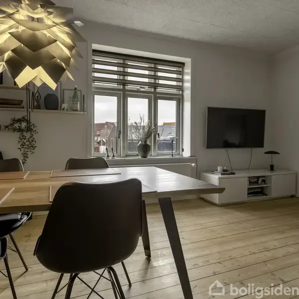 Et moderne stuerum med et stort spisebord, der står på et trægulv. Bordet er omgivet af stole. En lampe hænger over bordet. Der er en reol og et tv ved væggen.
