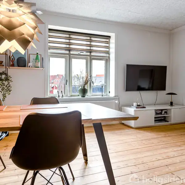 Spisebord med fire stole står i et lyst rum. Over bordet hænger en moderne lampe. Bagved ses en stor fladskærm og hylder med dekorationer. Gulvet er lyst træ.