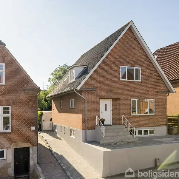 En rødstensvilla med spidst tag står mellem to lignende huse. Huset har hvide vinduesrammer og en hvid hoveddør. Solskin oplyser facaden, og fortovet er roligt.