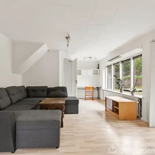 Grå sofa står i et lyst rum med trægulv ved store vinduer. Træbord og TV-bord er til stede, udgang til terrasse i højre side, udsigt til have.