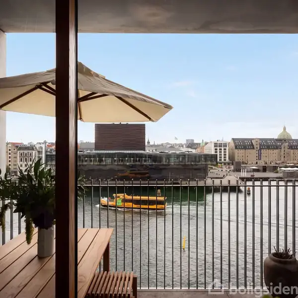 En altan har et træbord med planter og en parasol. Udsigt til en kanal med en gul båd og bygninger i baggrunden under en klar himmel.