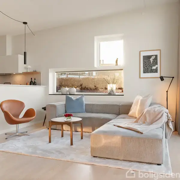 En lys stue med en beige hjørnesofa og et lille rundt bord foran. En brun lænestol står ved en halv væg til køkkenet. Store vinduer giver naturligt lysindfald.