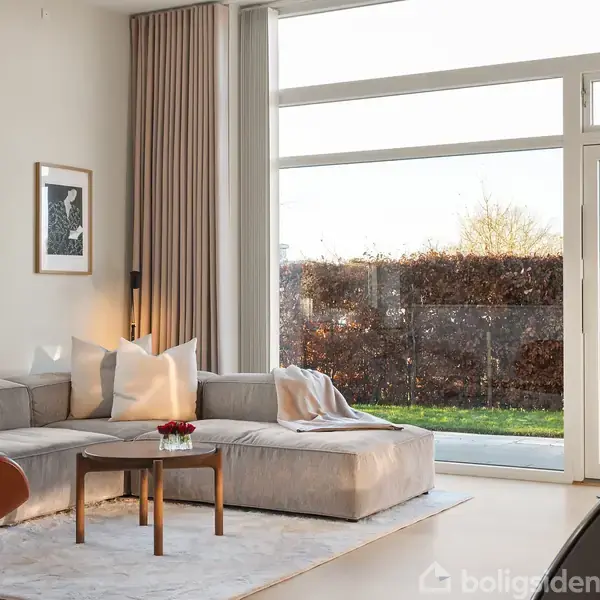 En lys stue har en grå hjørnesofa med puder, der står på et lyst tæppe. Et lille bord med blomster står foran sofaen. Store vinduer vender mod en have.