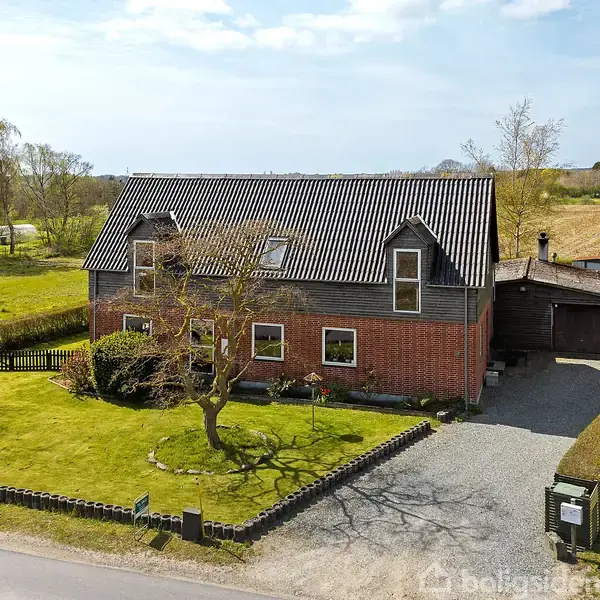 Et rødt murstenshus med sort tag står stille omgivet af en velplejet have og en grusvej. Huset har flere vinduer, og i baggrunden ses åbne marker og træer.