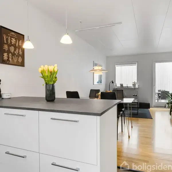 Køkkenø med hvide skuffer og en sort bordplade har en vase med gule tulipaner; spisestue med spisebord og stole i baggrunden. Væggene er hvide, gulvet er trægulv.