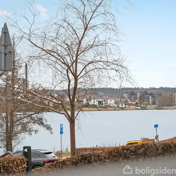 En bil holder parkeret ved en søbred omgivet af træer, buske og vejskilte. I baggrunden ses huse og en bakket landskabsudsigt under en blå himmel.