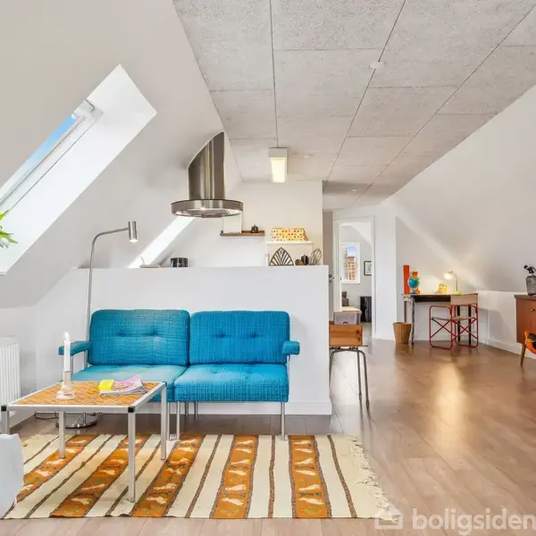 En blå sofa står ved et sofabord på et stribet tæppe i en stue. Rummet har skrå loftsvinduer, trælaminatgulv, og en blandet indretning med moderne og vintage møbler.