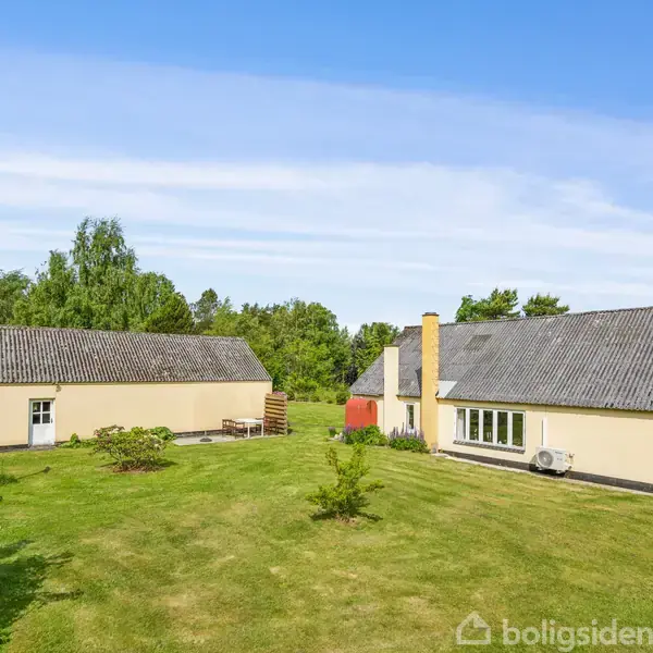 To gule bygninger står på en grøn græsplæne med spredte buske, omgivet af træer. Den ene bygning har en terrasse langs siden. Himmel er blå med lette skyer.