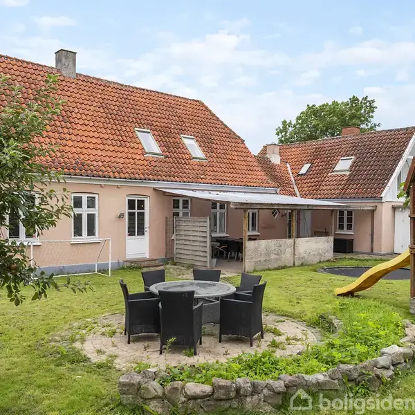 Et hus med rødtag ligger i en grøn have. Foran huset er der en rund terrasse med sorte havemøbler og en legekonstruktion med en rutsjebane.