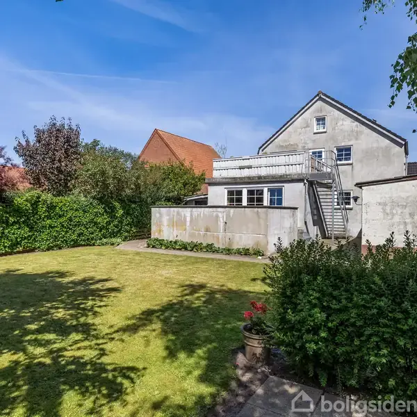 Et hus med baggård; huset har en trappe til en balkon, mens græsplænen er omkranset af buske og træer, hvilket skaber en hyggelig have.