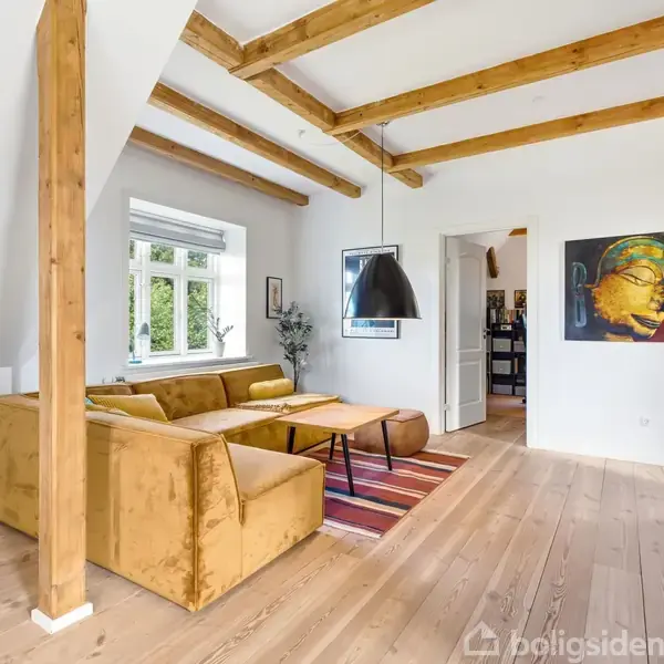En stue med en gul sofa ved et vindue. En sort lampe hænger over et bord med en stribet tæppe under. Til højre er en bogreol og et farverigt vægmaleri.