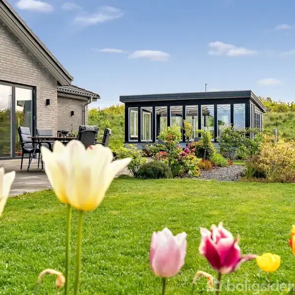 Hus med store vinduer står ved grøn have fyldt med farverige tulipaner; terrassen har et bord med stole. I baggrunden ses en moderne udestue omgivet af buske og græs.