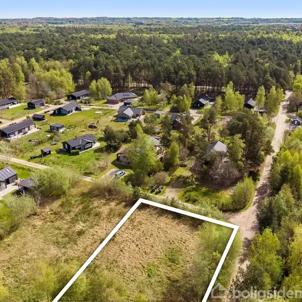 En afmærket, ubebygget grund ligger i et frodigt, skovklædt område med spredte huse og veje. Skoven strækker sig mod horisonten, omgivet af tæt beplantning og små græsplæner.
