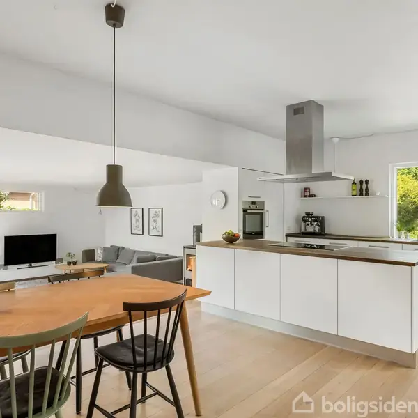 Et moderne køkken-alrum med en stor hvid køkkenø og indbyggede apparater. En spiseplads med træbord og stole står under pendellamper. Store vinduer giver udsigt til en have.