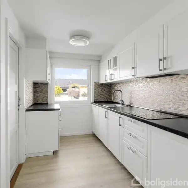 Et moderne køkken med hvide skabe og skuffer, sort bordplade, mosaik flisevæg. Et vindue i baggrunden giver udsigt til en lys dag udenfor. Ingen tekst i billedet.