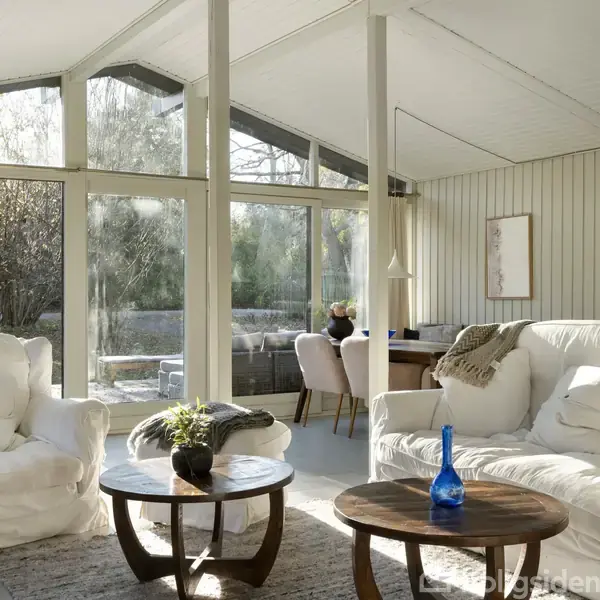 Et lyst stueområde med hvide møbler, herunder en sofa og lænestol, står i sollys tæt på store vinduer. Bordet har en blå flaske. Bakgrunden viser træer udenfor.