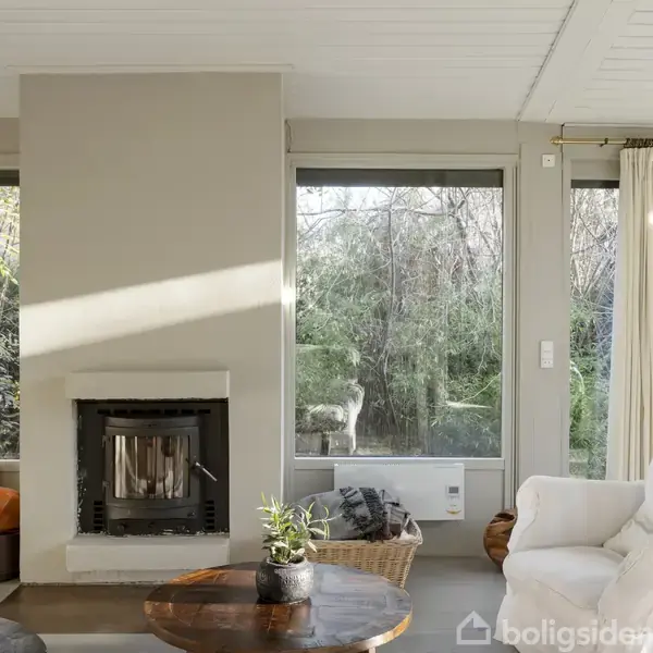 Pejs i stuen står stille ved en stor glasskydedør, der fører til en have med tæt bevoksning og en solstråle, der oplyser væggen. En hvid gardin hænger ved siden af døren.