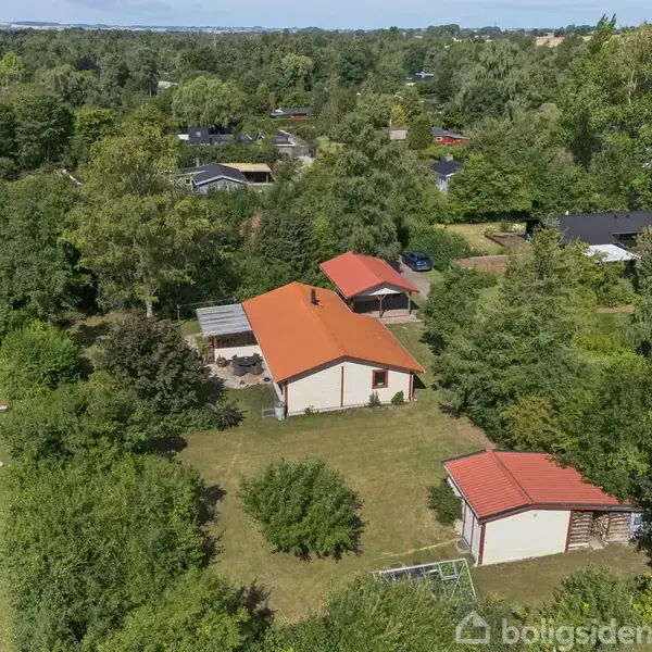 Et hus med rødt tag står i en grøn omgivelser af træer og buske. Græsplæne strækker sig omkring huset, som ligger tæt ved en vej.