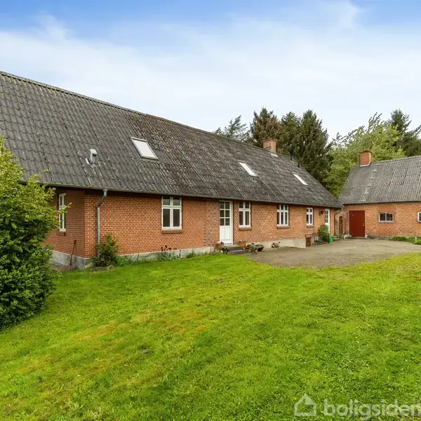 En rød murstensbygning med mørkt skråt tag står på en grøn græsplæne. Bygningen har flere vinduer og er omgivet af buske og træer under en blå himmel.