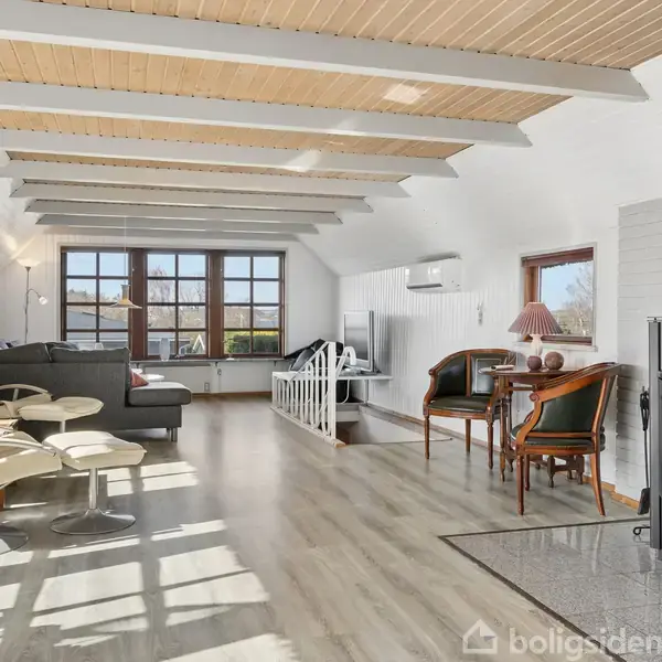 Stue med moderne møbler, hvor to lænestole og en sofa vender mod store vinduer. En brændeovn står til højre, og et spisebord med stole er bagved. Loftet har synlige bjælker.