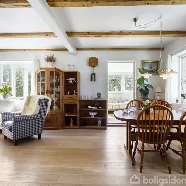 Stue med trægulv og synlige bjælker har en sofa, lænestol og spisebord. Skabe og dekorationer står langs væggene. Lyse vinduer slipper naturligt lys ind i rummet.