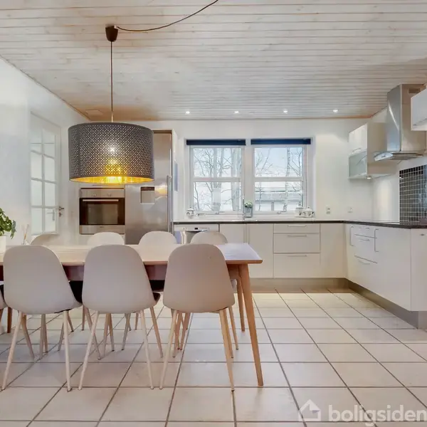 Et moderne køkken med hvidt bord og seks stole, hængende lampe ovenover. Vægur til venstre, stort vindue i baggrunden, hvide skabe og flisegulv.