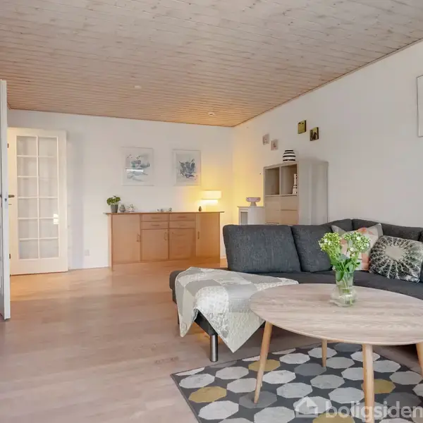 Stue med grå sofa og rundt træbord, dekoreret med blomster. Hylder og skab i baggrunden, belysning fra hjørnelampe og kunst på væggen. Lys trægulv.