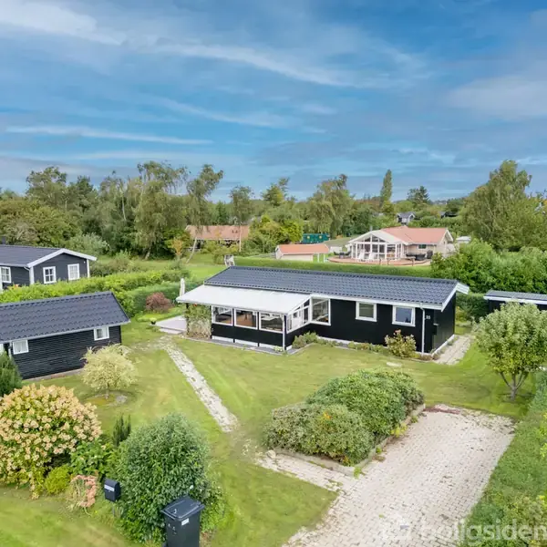 Sort sommerhus med veranda, omgivet af grønne plæner og træer i et villakvarter. Flere lignende huse ses i baggrunden, og en grusbelagt sti fører til huset.