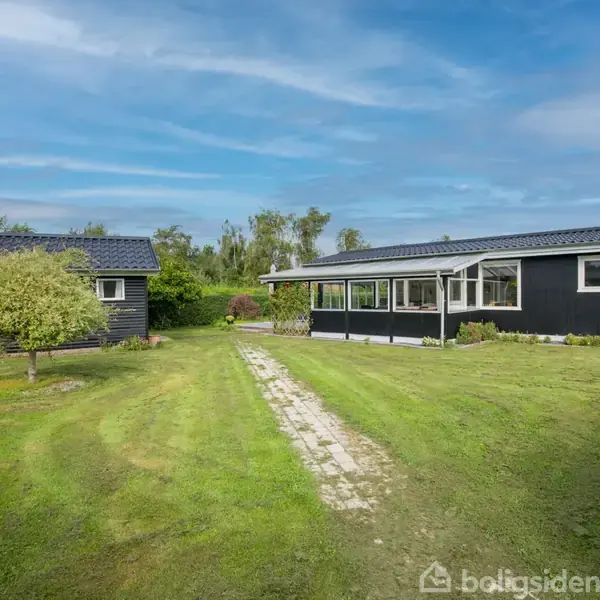 Et sortmalet hus og et skur står på en velholdt græsplæne under en blå himmel, omkranset af grønne buske og træer. En stensti forbinder bygningerne.