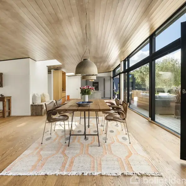 En stue med en grøn sofa og sofabord, åbner ud mod en spiseplads. Store glasvægge giver udsigt til en velplejet have. Trægulve og -loft skaber en varm atmosfære.