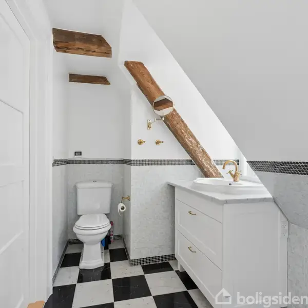 Toilet står ved siden af en skrå træbjælke i et lille badeværelse. Der er sort-hvidt ternet gulv og et hvidt håndvaskeskab til højre.