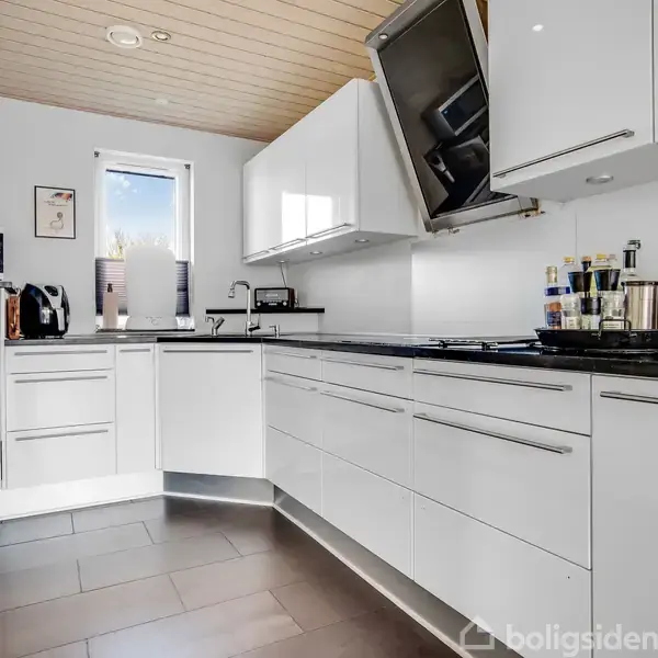 Hvidt, moderne køkken med indbyggede apparater og sort bordplade. Kedel og køkkenredskaber er synlige. Vinduet ved vasken slipper naturligt lys ind. Trælamineret loft og mørkt gulv.