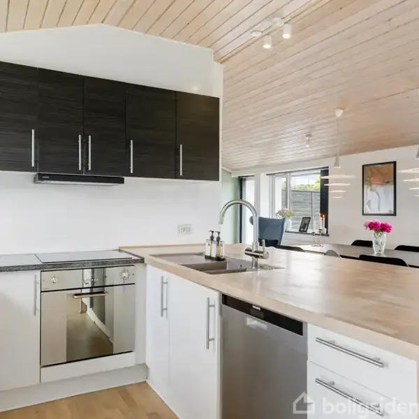 Køkken med hvide skabsfronter og sort overskab, køkkenø med vask og opvaskemaskine. Lys trælufter, spisebord med stole, vinduer og vase med lyserøde blomster i baggrunden.