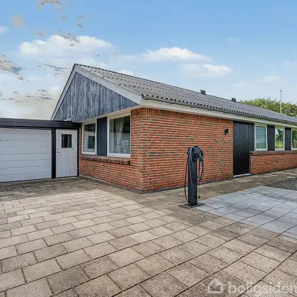 En murstensbungalow med sort tag og store vinduer står ved siden af en indbygget garage. Den er omgivet af en flisebelagt indkørsel og grønne buske i kanten af haven.