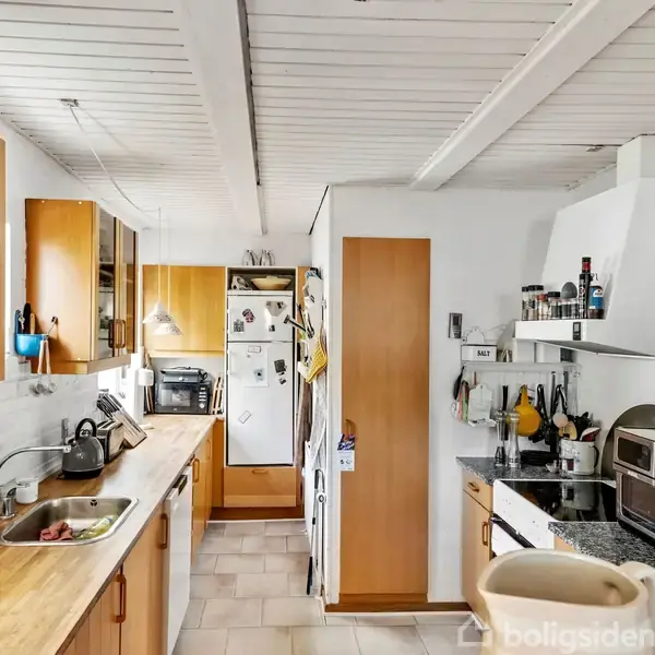 Et køkken med træskabe og -borde har en vask med køkkenredskaber ved siden af. Der er et køleskab dækket med magneter og en ovn med diverse madlavningstilbehør på bordet.