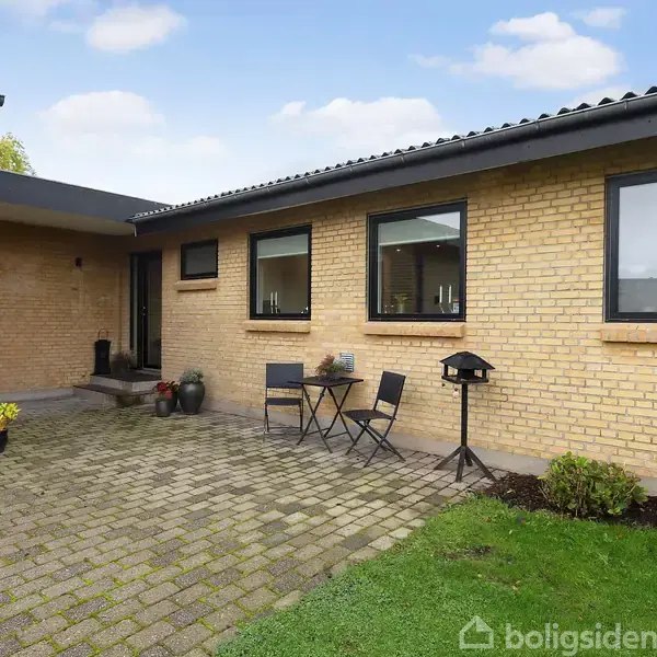 En gårdhave med terrasse i en gul murstensbygning. Der står et cafébord med to sorte stole samt potteplanter nær vinduerne. Flisebelægning omgiver området.